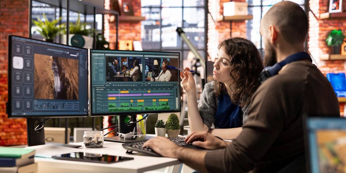Two professionals collaborating at a desk with dual monitors displaying video editing software, timelines, and media assets, representing teamwork, planning, and digital workflow optimization during a Business Central Migration project.