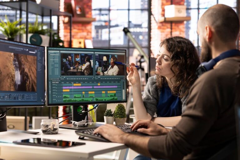 Two professionals collaborating at a desk with dual monitors displaying video editing software, timelines, and media assets, representing teamwork, planning, and digital workflow optimization during a Business Central Migration project.
